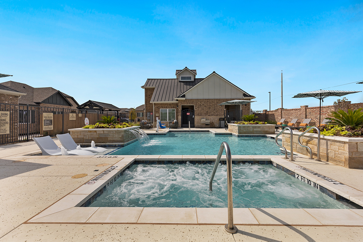 A hot tub with bubbling water is in the foreground, leading to a rectangular outdoor pool with lounge chairs. A small building and clear blue sky are in the background.