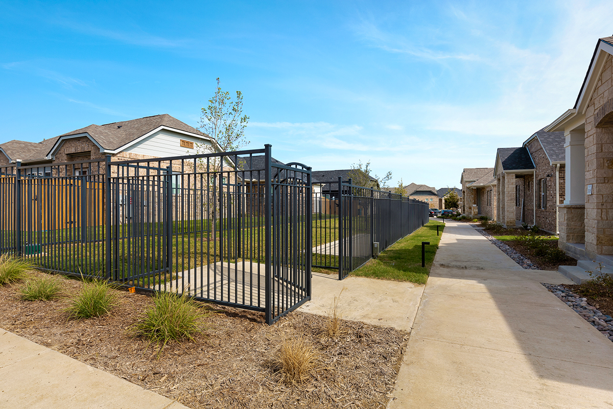 A sidewalk runs beside a fenced grassy yard between two rows of modern houses under a clear blue sky.