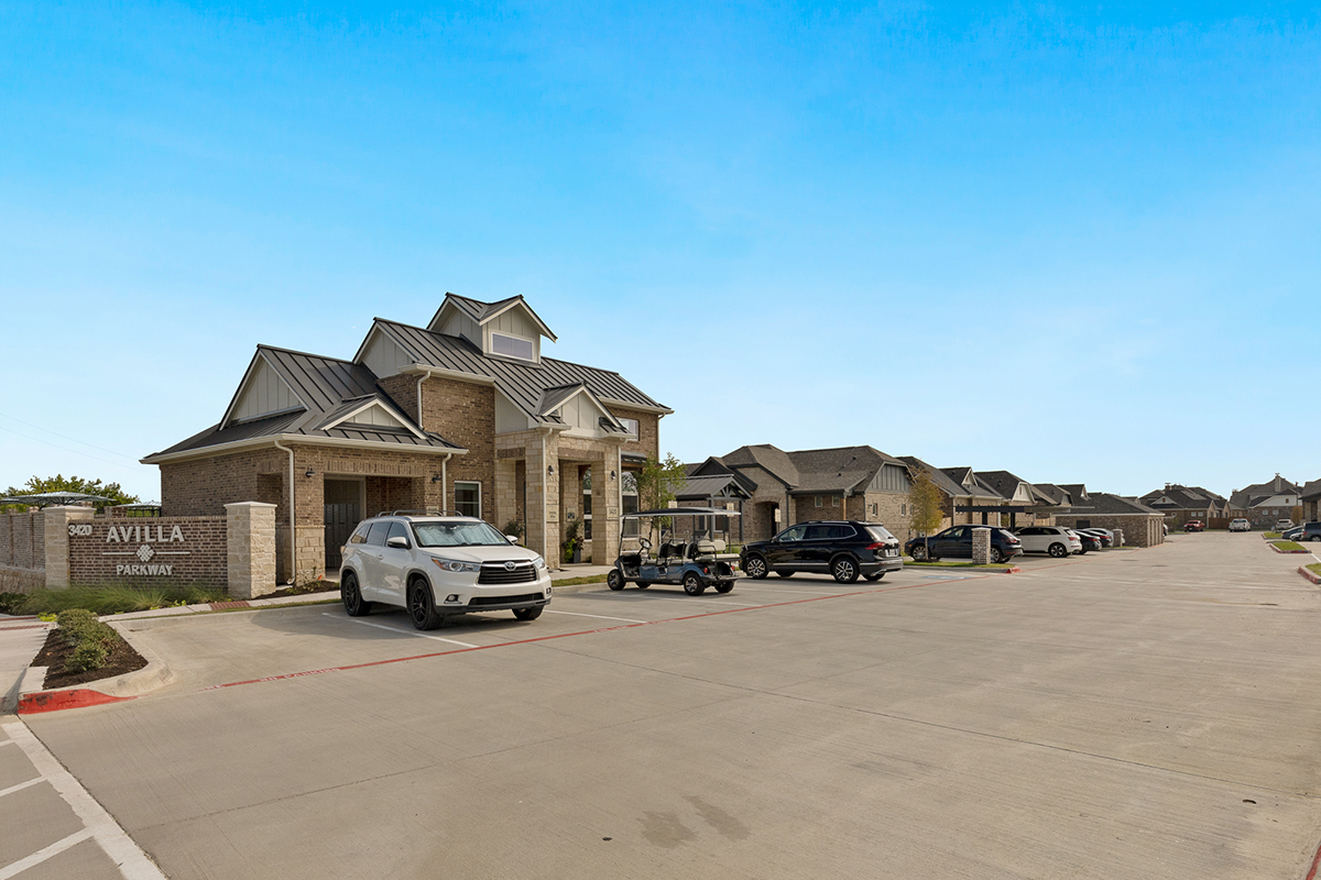 A brick building with a sign reading Avilla Parkway, several parked cars, and a golf cart are visible in front. Single-story homes line the right side under a clear blue sky.