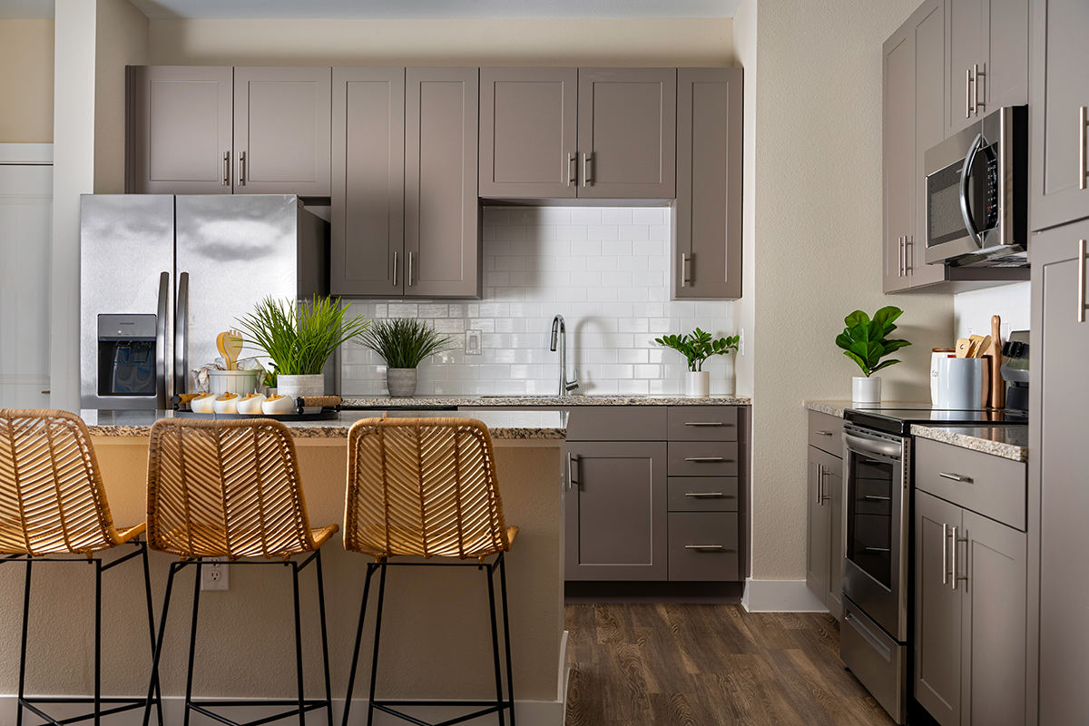 Modern kitchen with gray cabinets, stainless steel appliances, a large island with three woven stools, potted plants, and white subway tile backsplash. The floor is wood, and the space is well-lit and tidy.