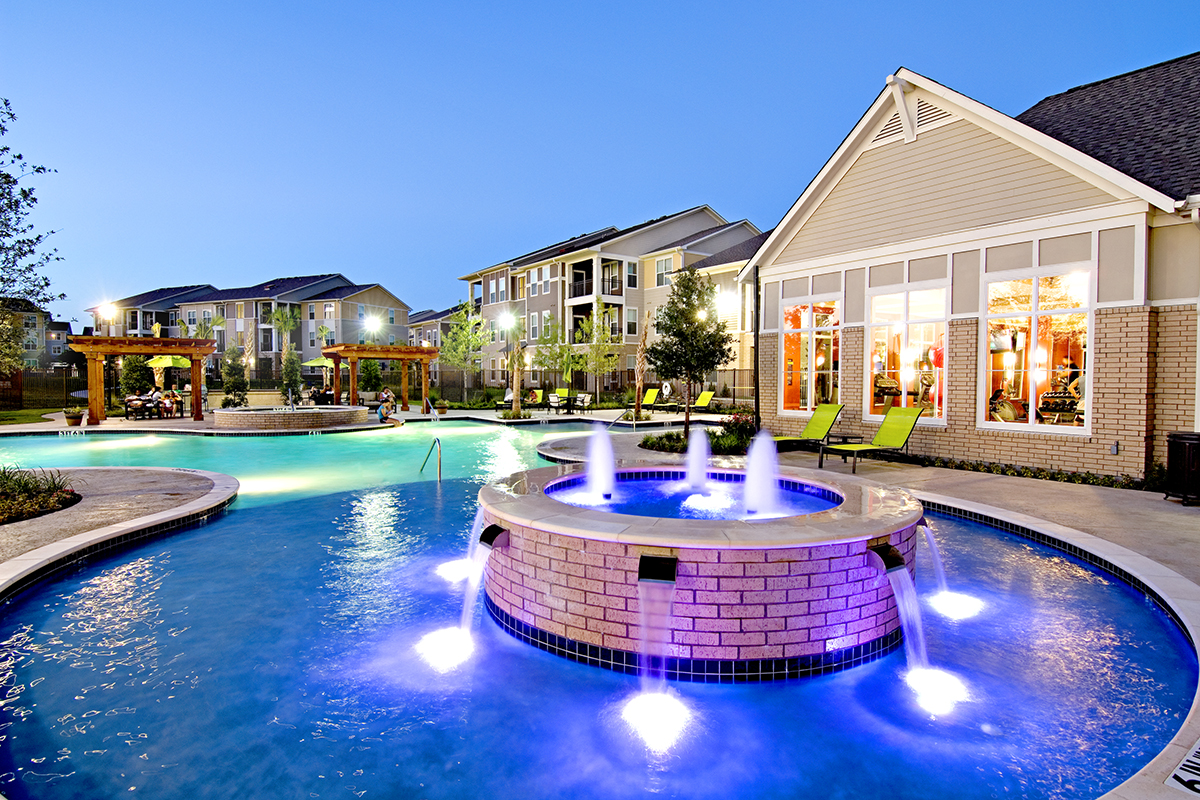 A modern apartment complex at dusk features a glowing pool with water fountains, surrounded by lounge chairs, greenery, and well-lit buildings with large windows.