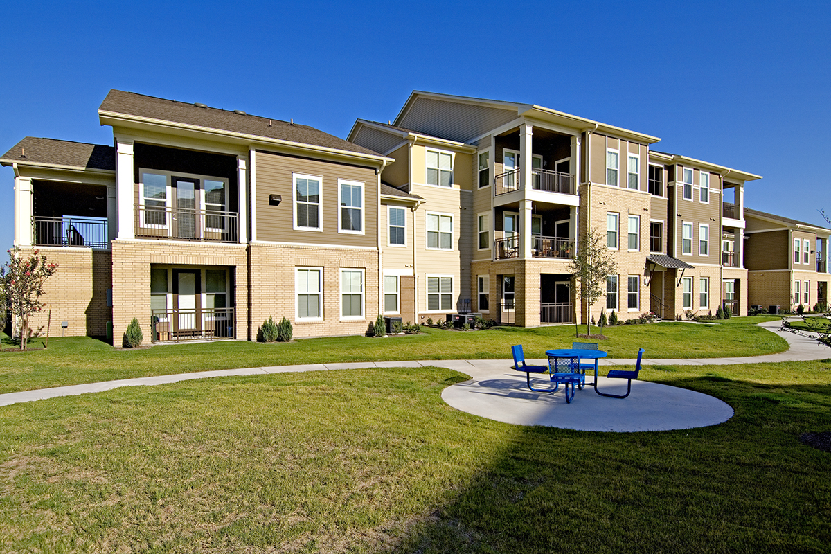 Three-story apartment building with balconies and large windows, surrounded by well-kept grass. A blue picnic table with benches sits on a concrete pad along a curving sidewalk under a clear blue sky.