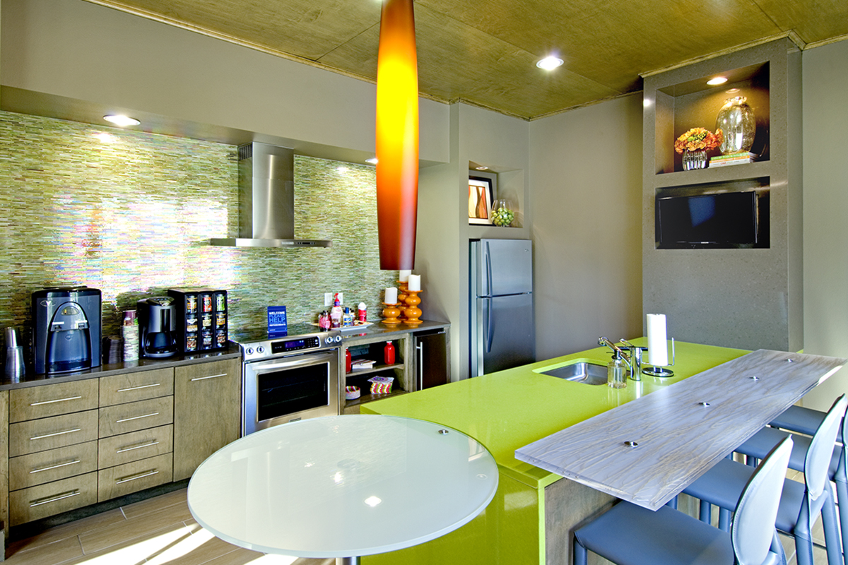 Modern kitchen with green island, bar stools, stainless steel appliances, coffee machines, glass backsplash, a wall-mounted TV, and decorative shelves. Bright pendant light hangs over the island.