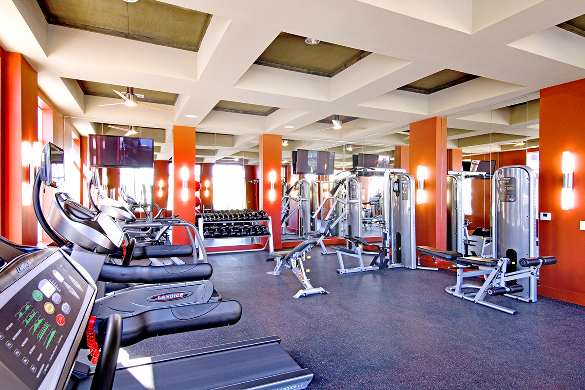 Bright gym with treadmills, weight machines, free weights, mirrors, orange walls, and ceiling lights. TVs are mounted on the walls above the equipment. The space is clean and well-lit.