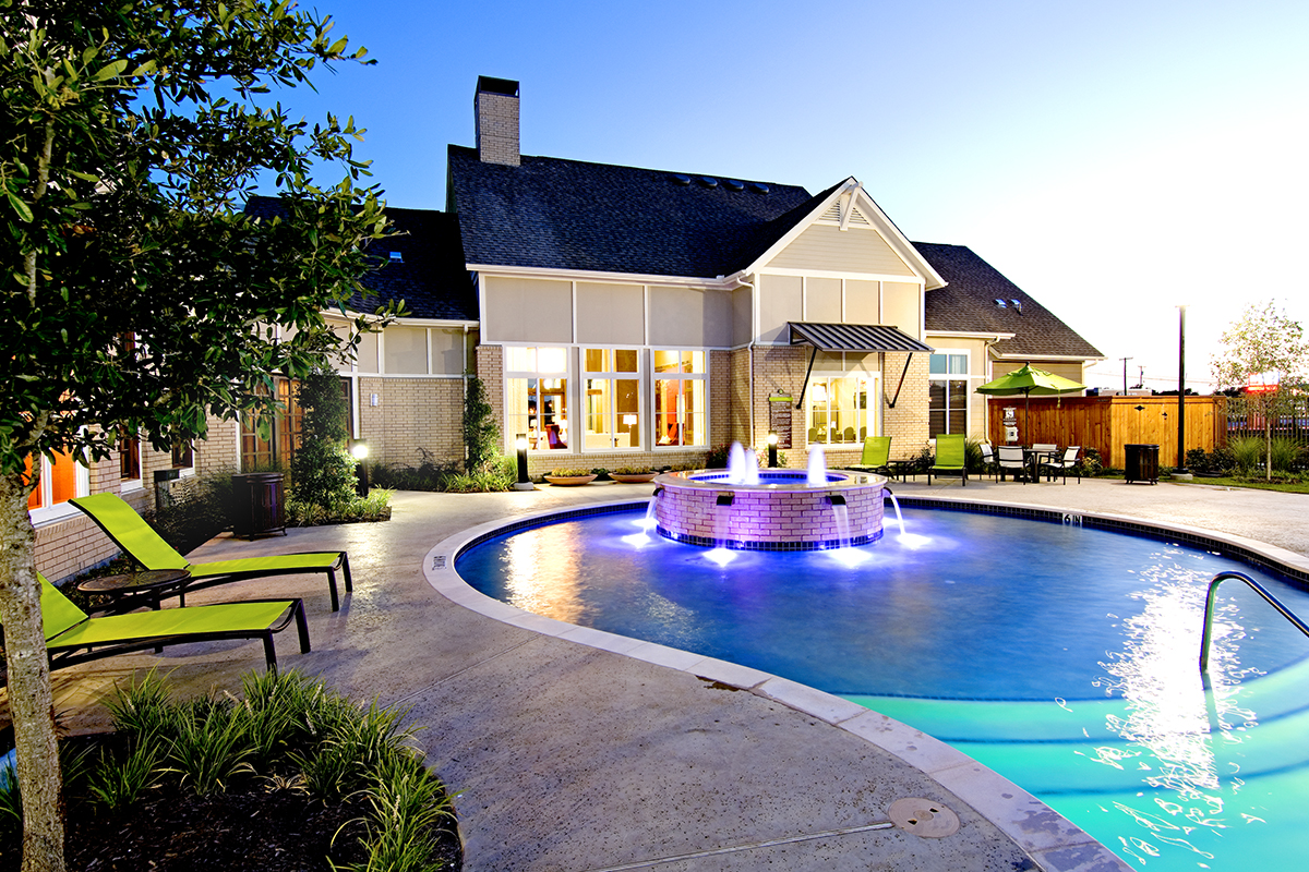 A modern house with large windows overlooks a lit swimming pool featuring a circular fountain. Green lounge chairs sit on the poolside patio at dusk.