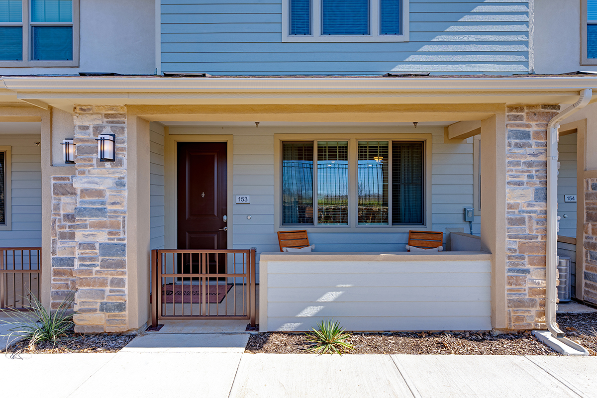 A townhouse exterior with a brown front door, stone and siding facade, a small fenced patio, two wooden chairs, and a window with closed blinds.