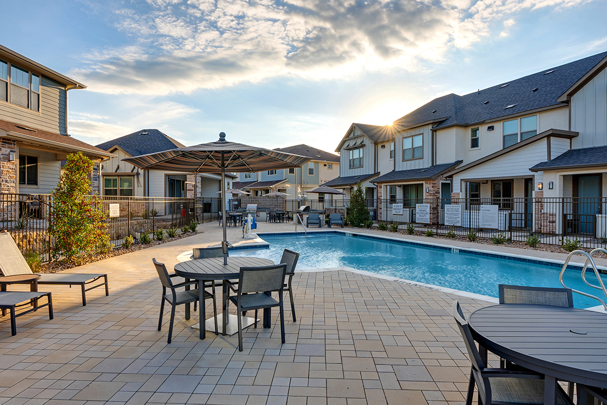 A fenced outdoor swimming pool with lounge chairs, tables, and umbrellas, surrounded by modern townhouses under a partly cloudy sky at sunset.