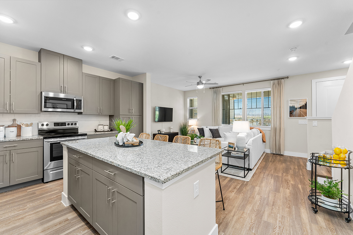 Modern open-concept kitchen and living room with gray cabinets, granite countertops, stainless steel appliances, wood floors, barstools, and a seating area with a sofa, TV, and large windows letting in natural light.
