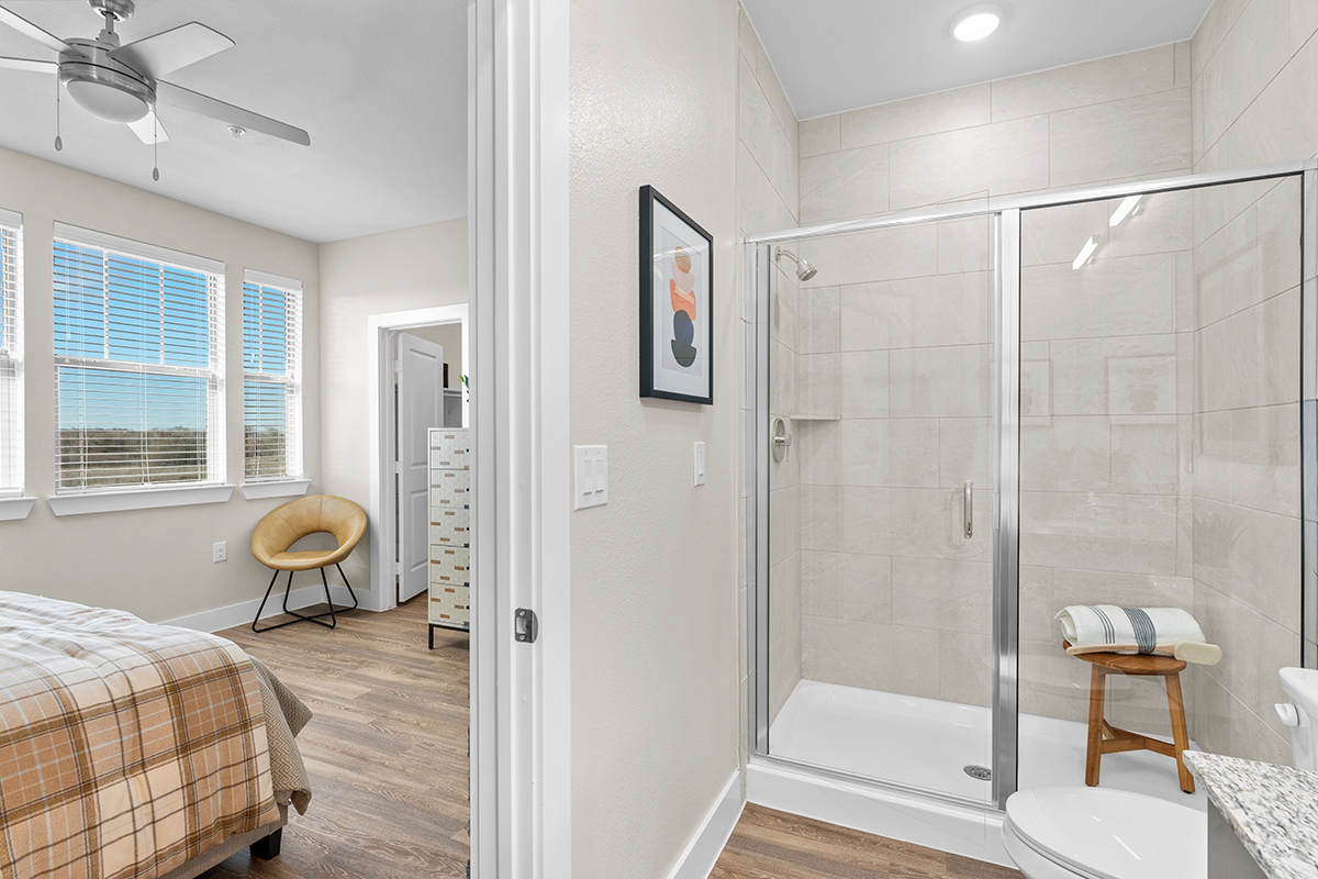A modern bedroom with wood flooring is adjacent to a bright bathroom featuring a glass-enclosed shower, beige tiles, a wooden stool with towels, and a simple wall art piece.