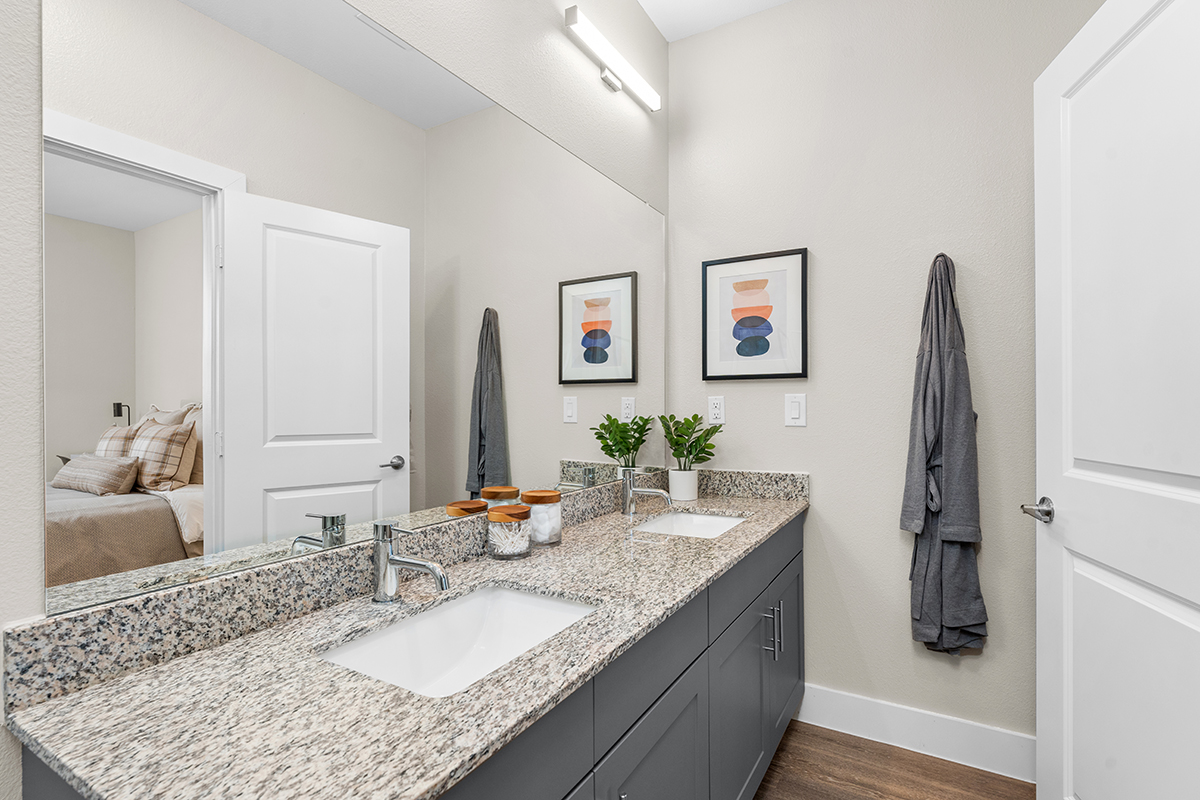 Modern bathroom with granite countertop, dual sinks, dark gray cabinets, two framed abstract prints, decorative jars, a plant, and two robes hanging on the wall. A bedroom is visible through the open door.