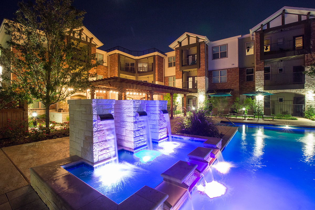 Night view of an apartment complex courtyard with illuminated stone water fountains and a lit swimming pool, surrounded by landscaped greenery and multi-story buildings.