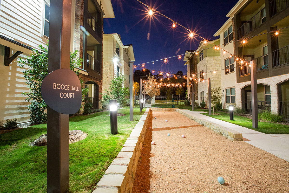 A bocce ball court at night, illuminated by string lights, is surrounded by modern apartment buildings and green lawns. Several bocce balls are scattered on the gravel court.
