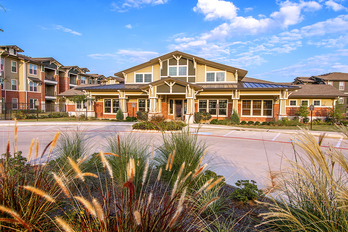 A modern apartment complex with a central building, surrounded by landscaped grasses and a mostly empty parking lot under a bright, partly cloudy sky.