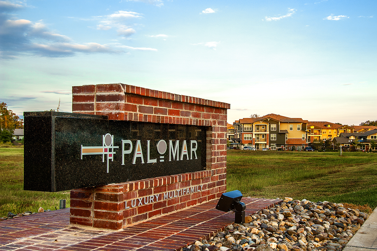 A brick and stone sign for PALOMAR Luxury Multifamily stands on a landscaped corner, with modern townhouses and a grassy field in the background under a blue sky.