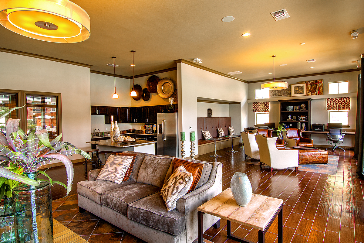 Modern open living space with brown wood floors, a sofa with patterned pillows, kitchen area, dining table, and a cozy sitting area; decor includes vases, plants, and wall art. Warm lighting creates a welcoming atmosphere.