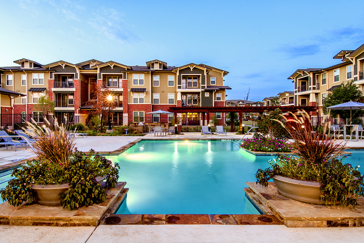 A large, inviting swimming pool surrounded by lush planters, lounge chairs, and three-story apartment buildings under a bright blue sky at sunset.