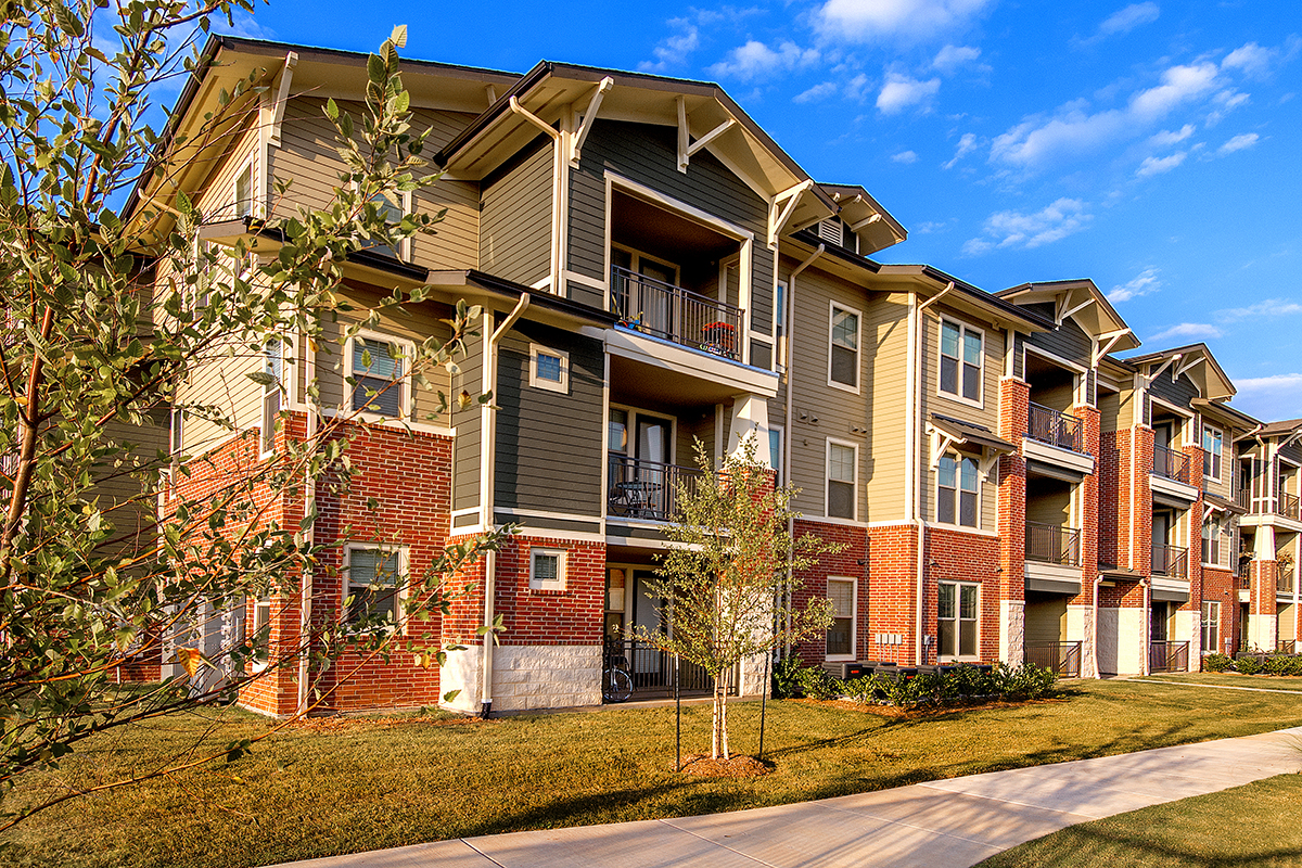 Modern three-story apartment building with red brick and beige siding, balconies, and large windows, surrounded by grass, small trees, and a sidewalk under a blue sky with scattered clouds.