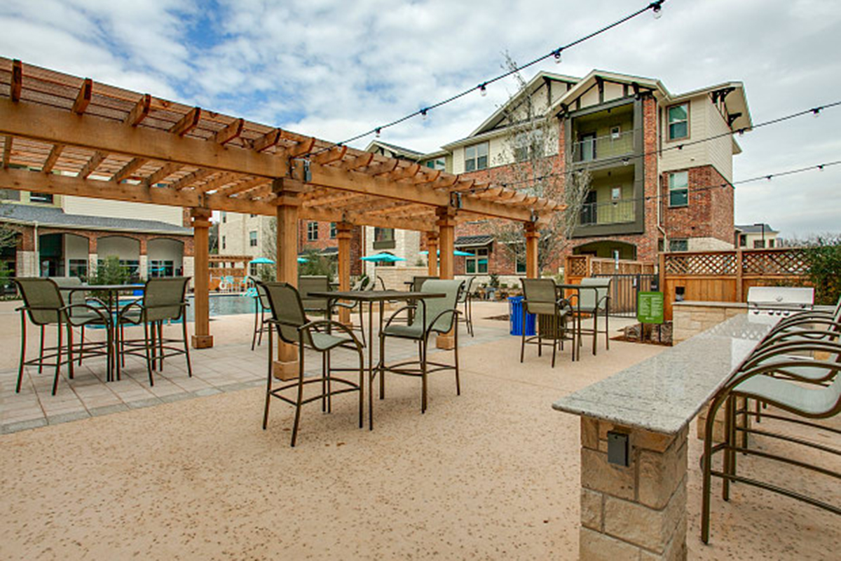 Outdoor patio area with a wooden pergola, tall tables and chairs, string lights overhead, and apartment buildings in the background.