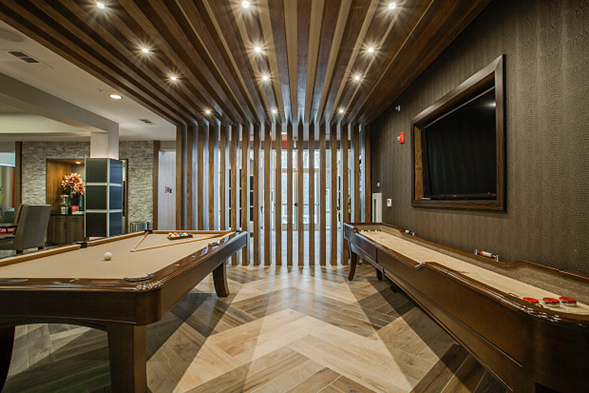 A modern game room with a pool table and shuffleboard, wood-paneled ceiling, chevron-patterned floor, and a large wall-mounted TV. Glass doors and slatted wood dividers let in natural light.